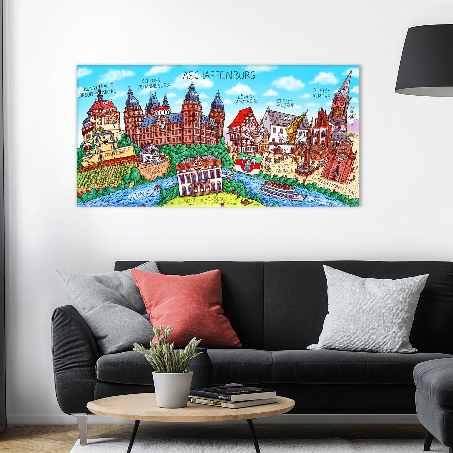 Jacques Tilly Aschaffenburg Stadtbild auf Leinwand mit Holzrahmen über einem schwarzen Sofa mit einem roten und zwei grauen Kissen und einem runden Couchtisch aus Holz davor.