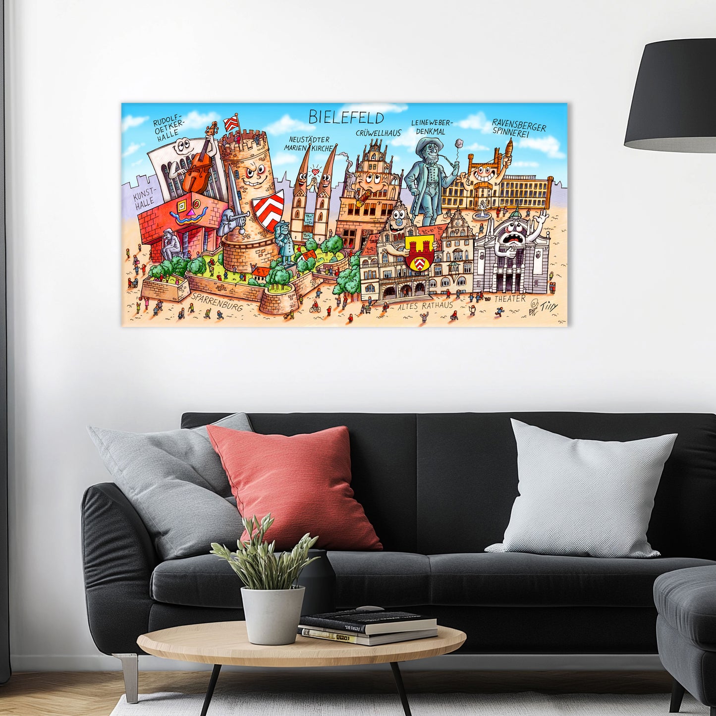 Jacques Tilly Bielefeld Stadtbild als Acrylbild mit Alu-Rückrahmen über einem schwarzen Sofa mit einem roten und zwei grauen Kissen und einem runden Couchtisch aus Holz davor.