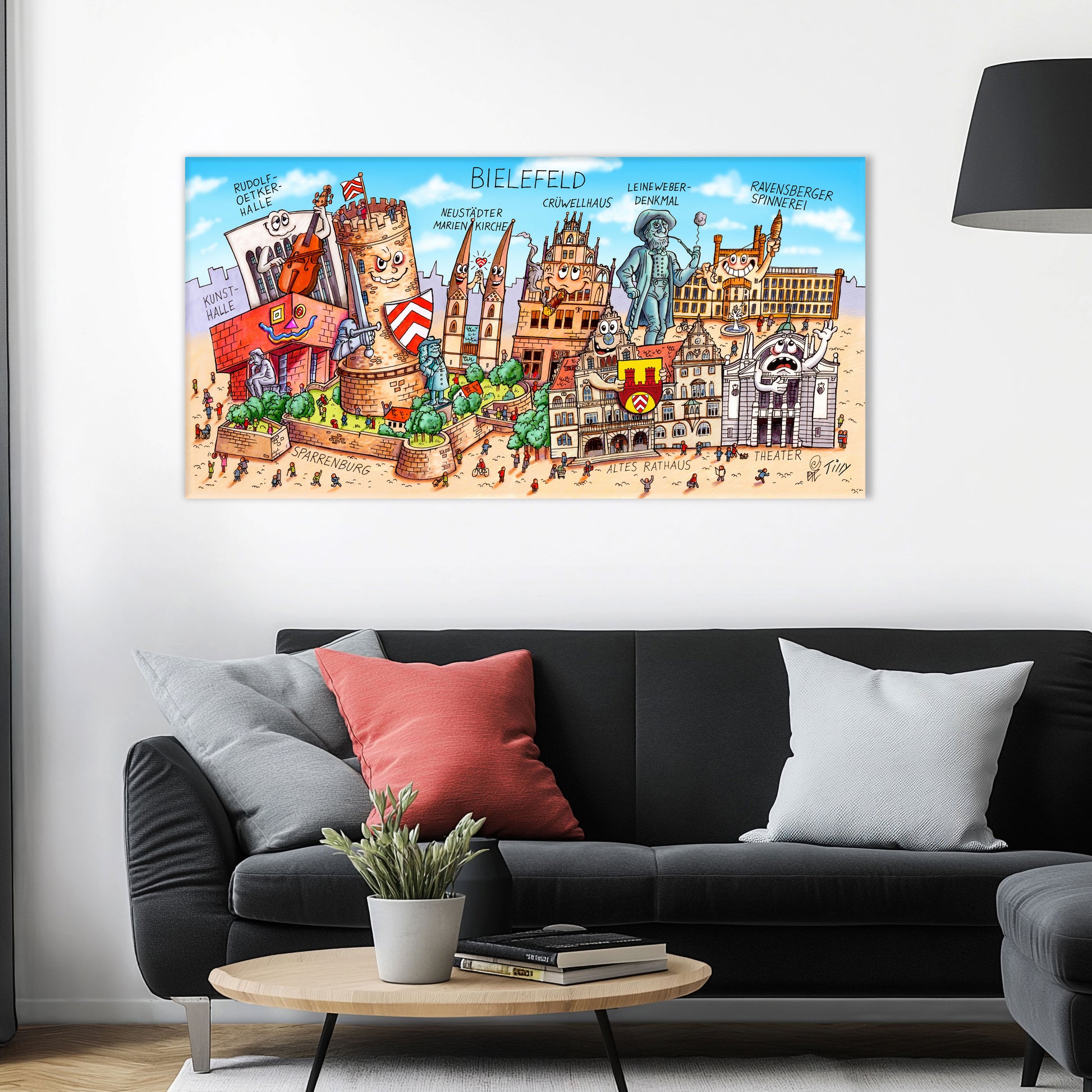 Jacques Tilly Bielefeld Stadtbild als Acrylbild mit Alu-Rückrahmen über einem schwarzen Sofa mit einem roten und zwei grauen Kissen und einem runden Couchtisch aus Holz davor.