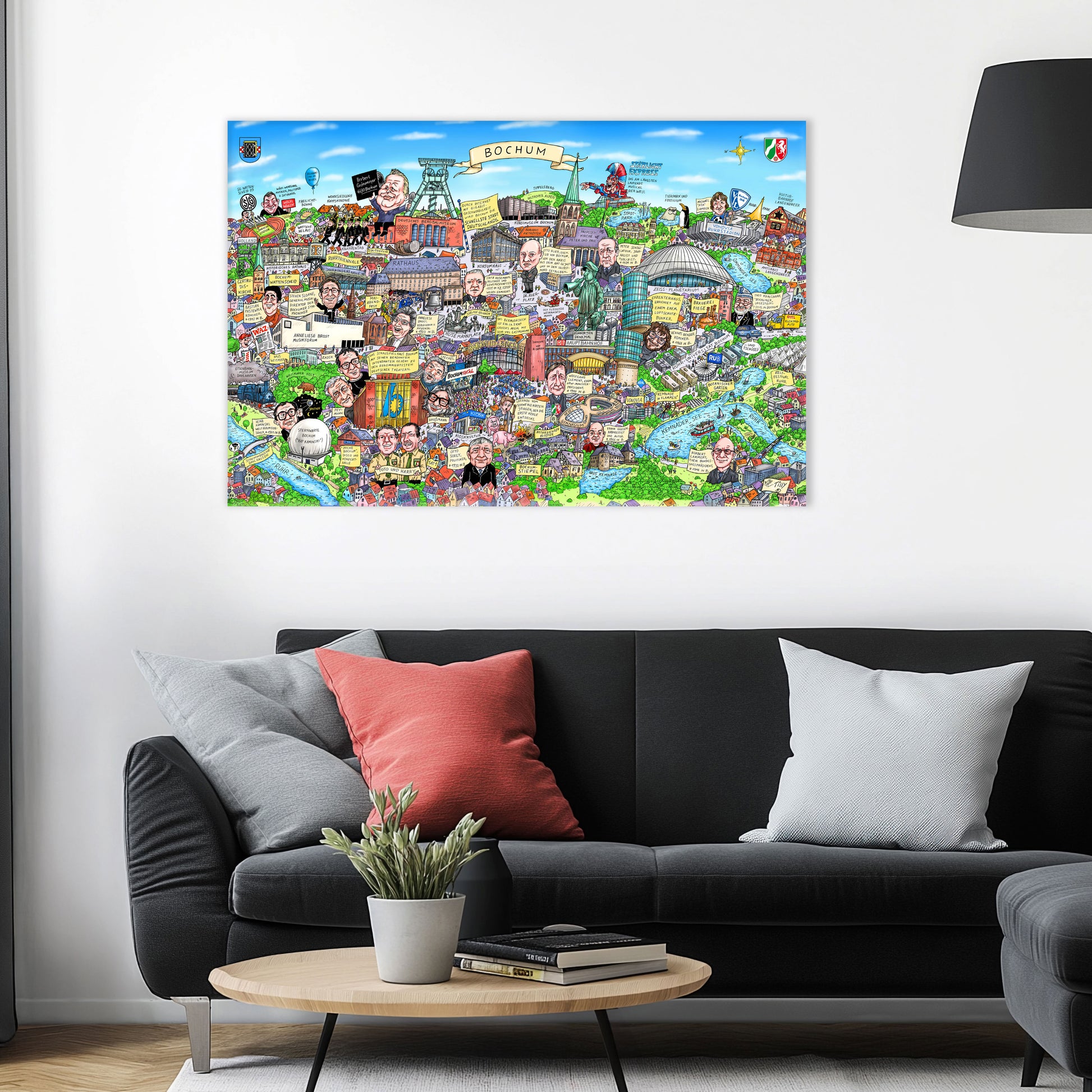 Jacques Tilly Bochum Wimmelbild Stadtbild als Acrylbild mit Alu-Rückrahmen über einem schwarzen Sofa mit einem roten und zwei grauen Kissen und einem runden Couchtisch aus Holz davor.