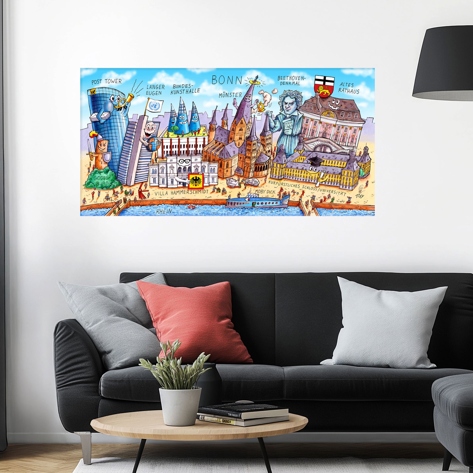 Jacques Tilly Bonn Stadtbild auf Leinwand mit Holzrahmen über einem schwarzen Sofa mit einem roten und zwei grauen Kissen und einem runden Couchtisch aus Holz davor.