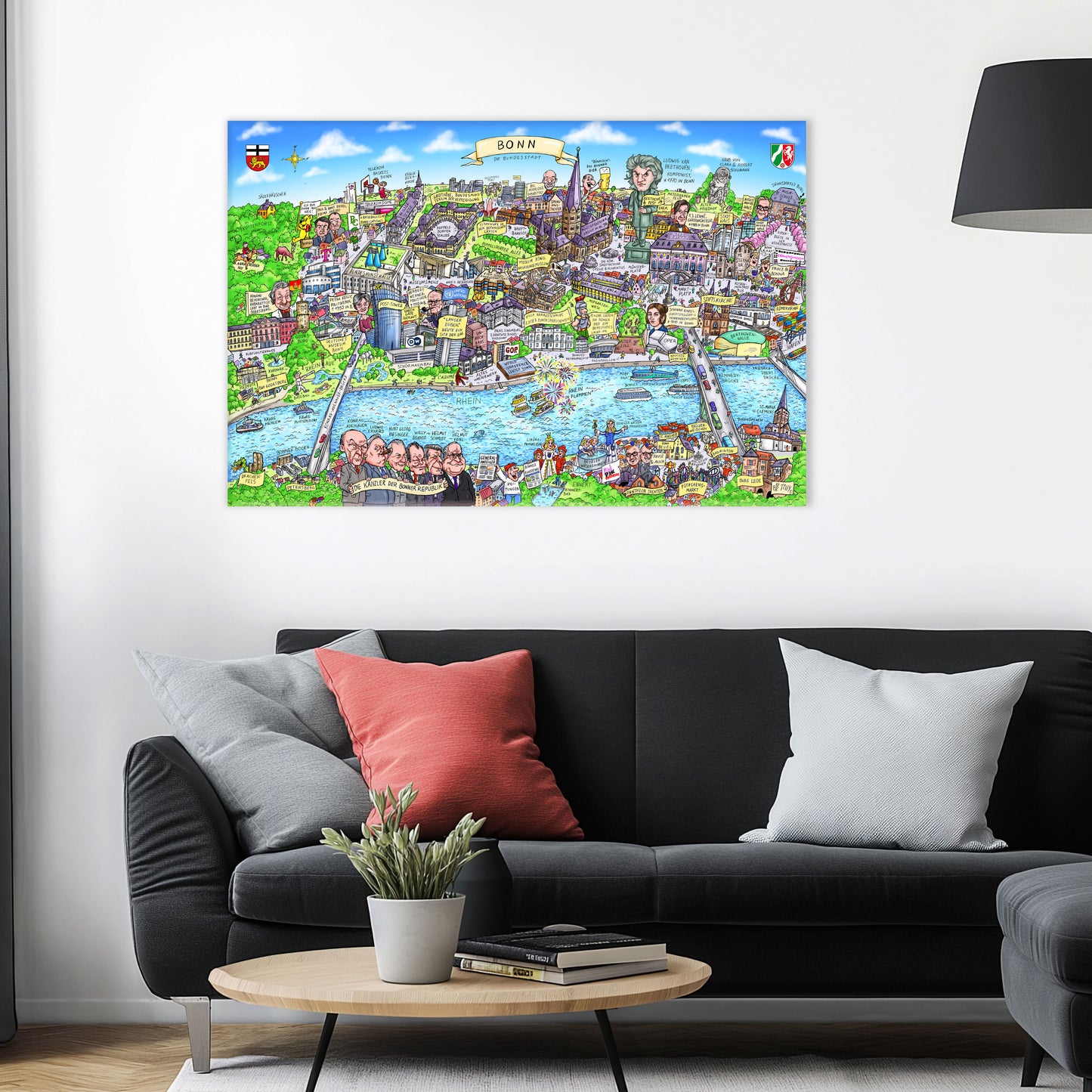 Jacques Tilly Bonn Stadtbild als Acrylbild mit Alu-Rückrahmen über einem schwarzen Sofa mit einem roten und zwei grauen Kissen und einem runden Couchtisch aus Holz davor.