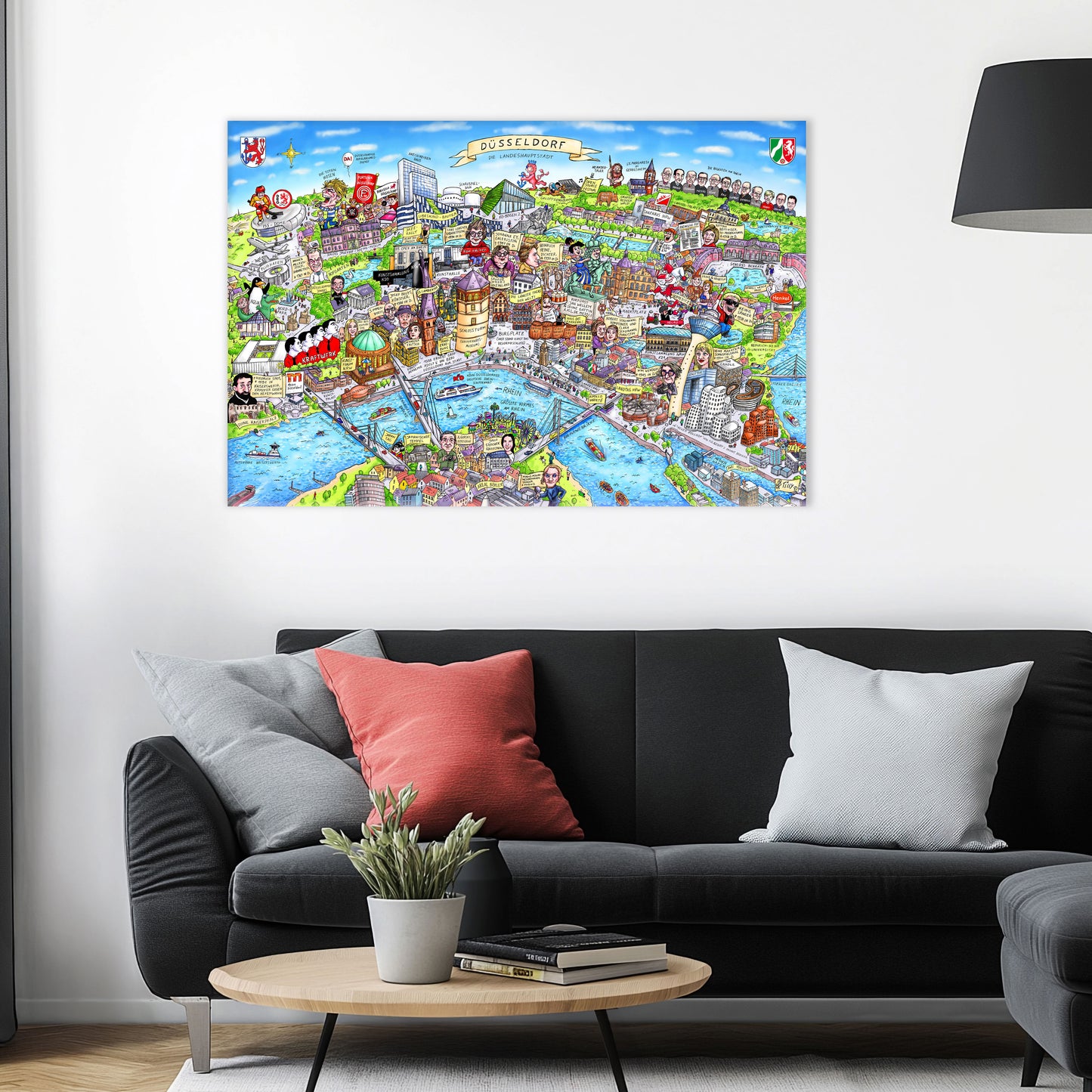 Jacques Tilly Düsseldorf Wimmelbild als Acrylbild mit Alu-Rückrahmen über einem schwarzen Sofa mit einem roten und zwei grauen Kissen und einem runden Couchtisch aus Holz davor.