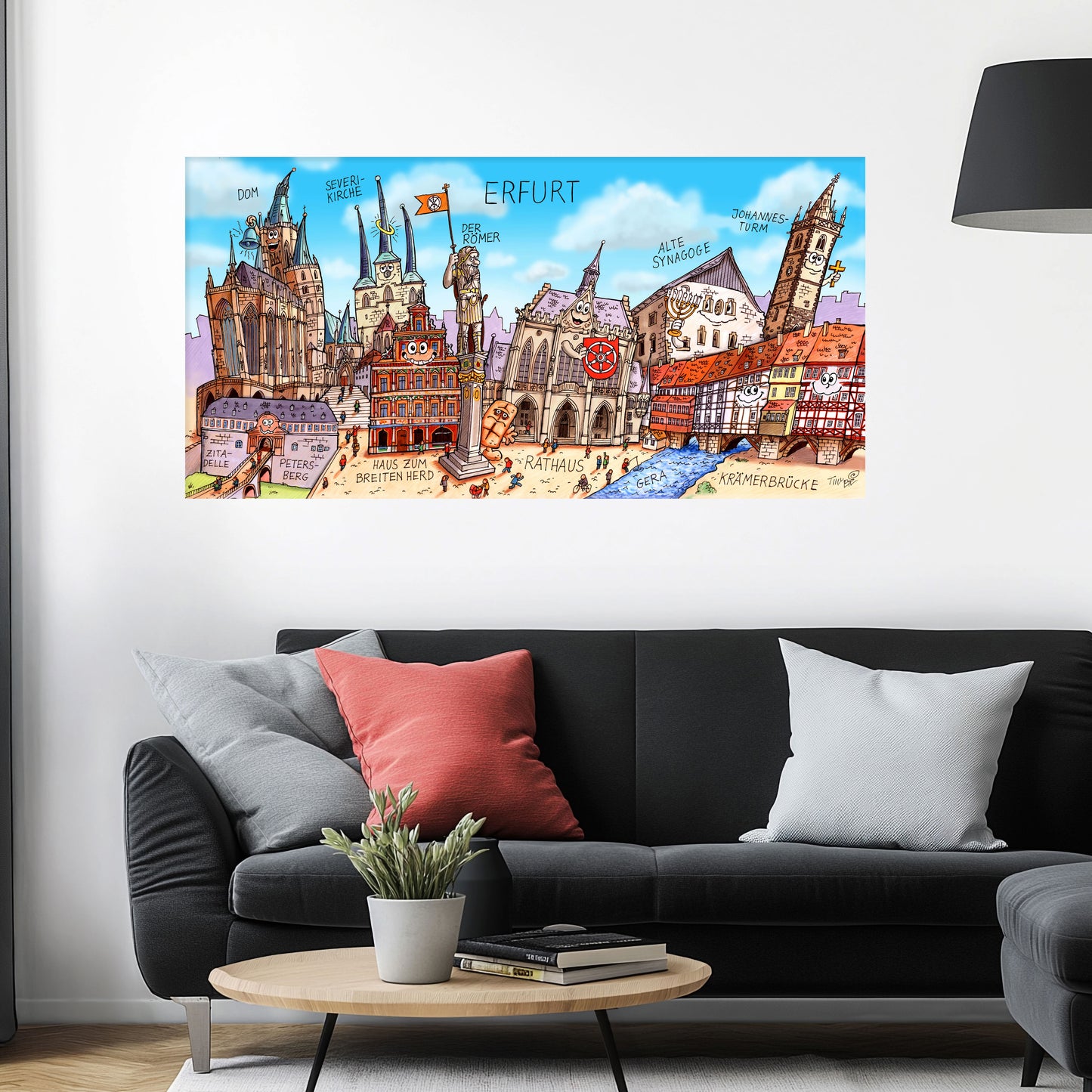 Bild an der Wand mit dem Stadtbild Erfurt von Jacques Tilly über einem schwarzen Sofa mit einem roten und zwei grauen Kissen und einem runden Couchtisch aus Holz davor.