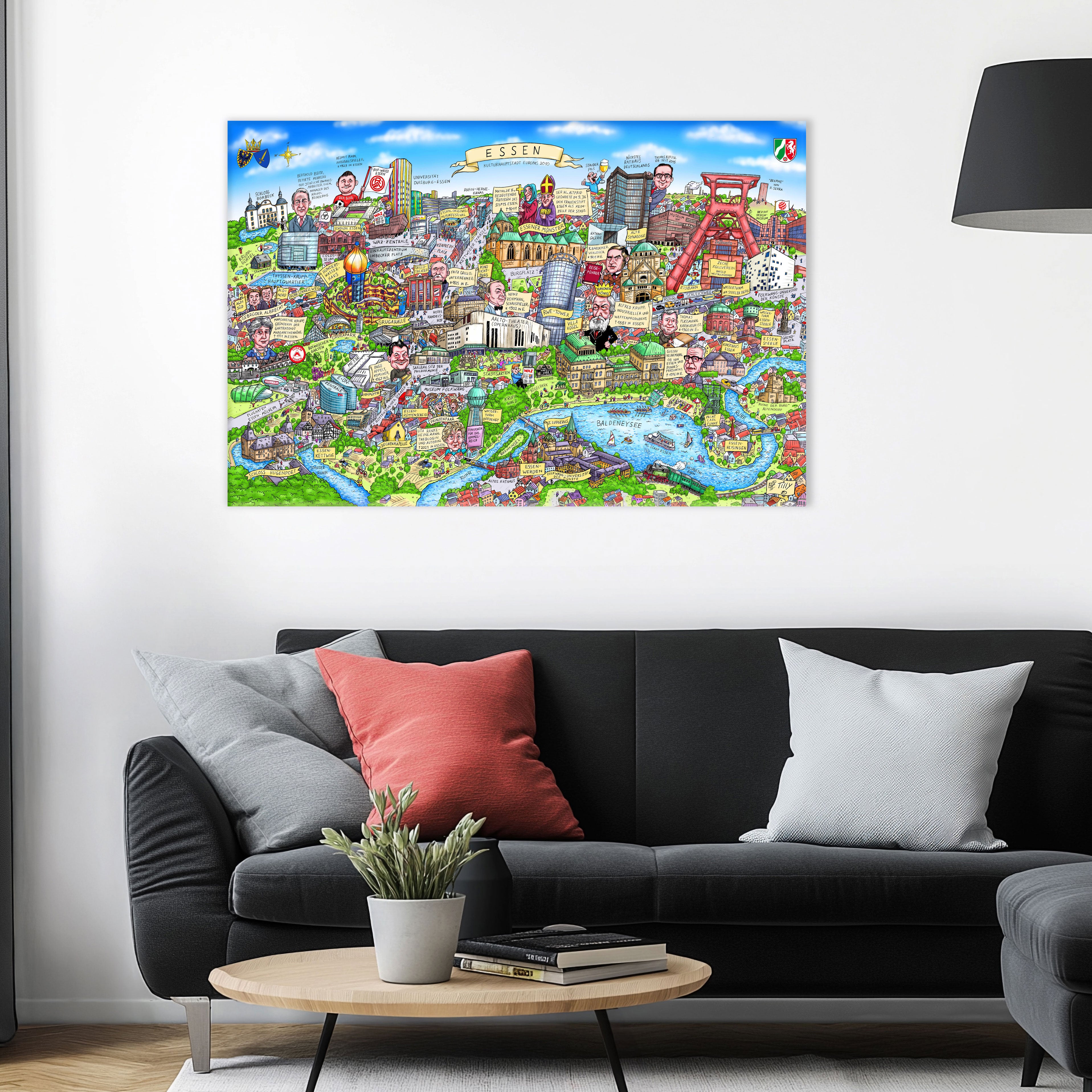 Jacques Tilly Essen Stadtbild als Acrylbild mit Alu-Rückrahmen über einem schwarzen Sofa mit einem roten und zwei grauen Kissen und einem runden Couchtisch aus Holz davor.