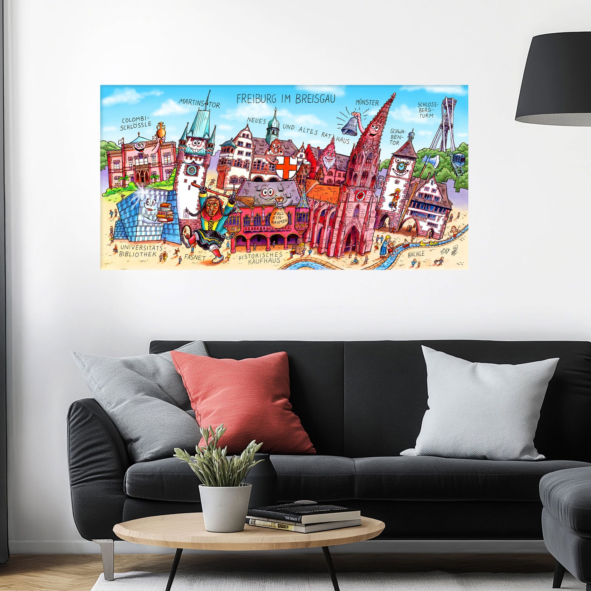 Jacques Tilly Freiburg Stadtbild als Acrylbild mit Alu-Rückrahmen über einem schwarzen Sofa mit einem roten und zwei grauen Kissen und einem runden Couchtisch aus Holz davor.