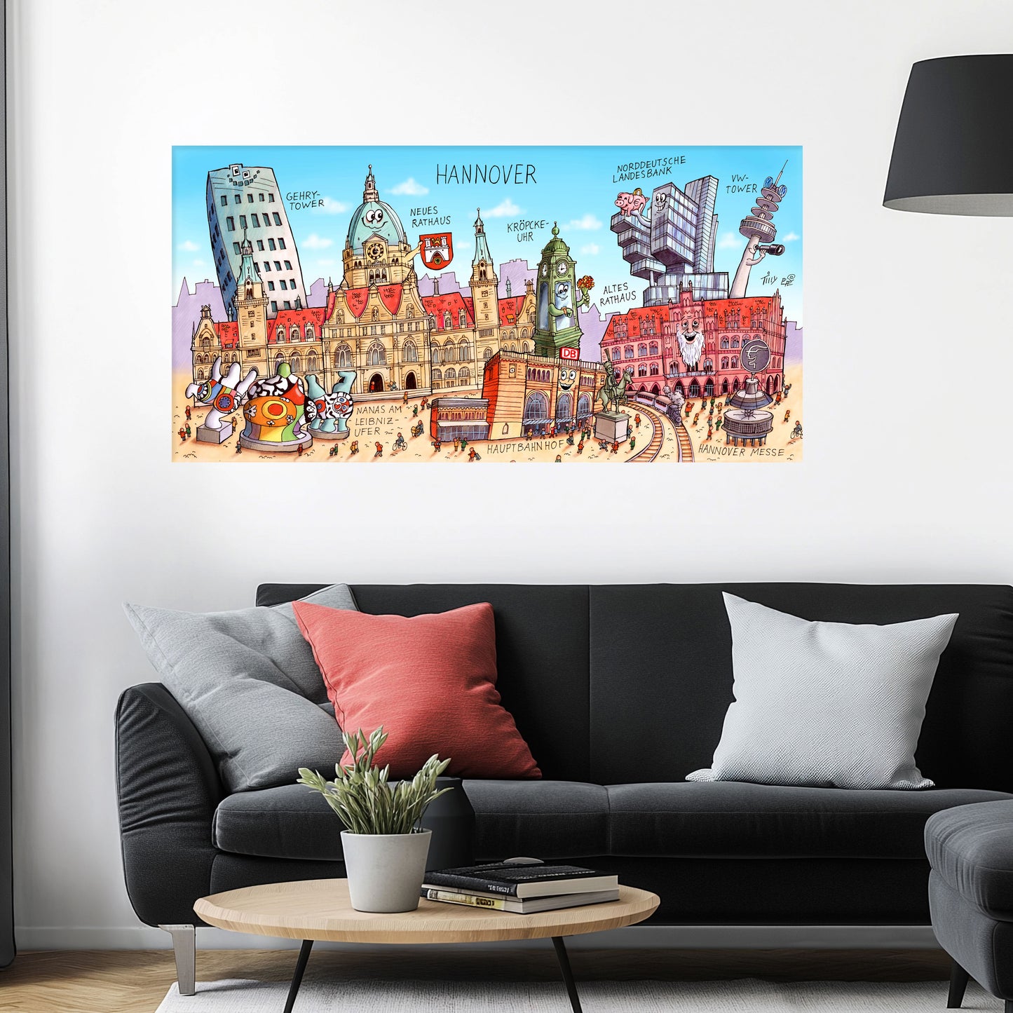 Jacques Tilly Hannover Stadtbild als Acrylbild mit Alu-Rückrahmen über einem schwarzen Sofa mit einem roten und zwei grauen Kissen und einem runden Couchtisch aus Holz davor.