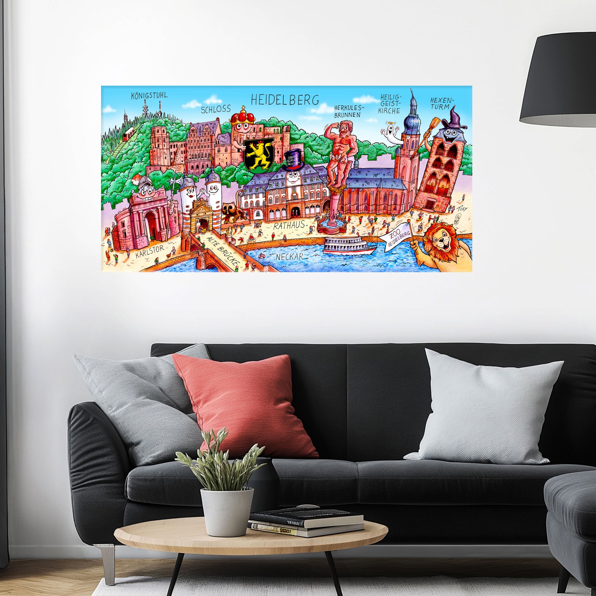 Jacques Tilly Heidelberg Stadtbild auf Leinwand mit Holzrahmen über einem schwarzen Sofa mit einem roten und zwei grauen Kissen und einem runden Couchtisch aus Holz davor.