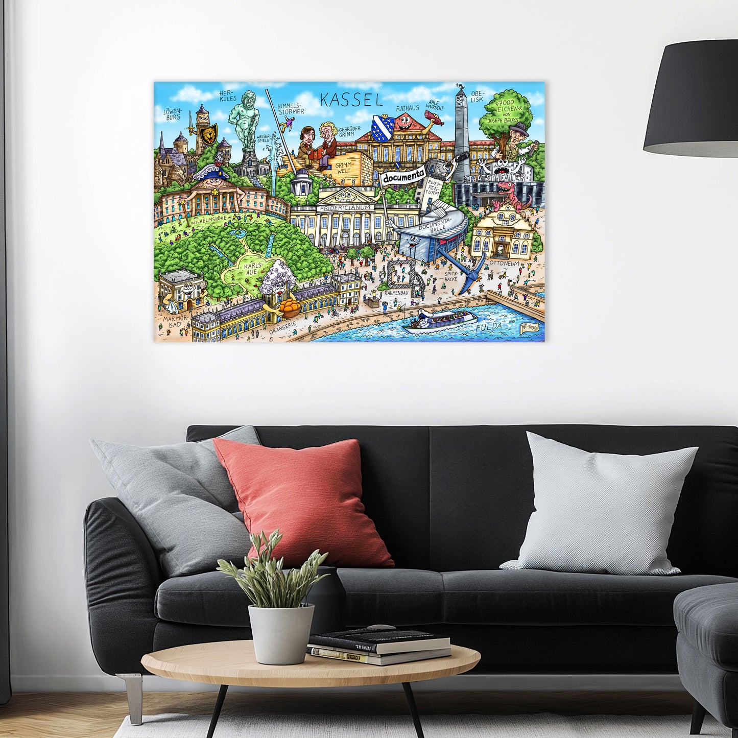 Jacques Tilly Kassel Stadtbild Wimmelbild auf Leinwand mit Holzrahmen über einem schwarzen Sofa mit einem roten und zwei grauen Kissen und einem runden Couchtisch aus Holz davor.