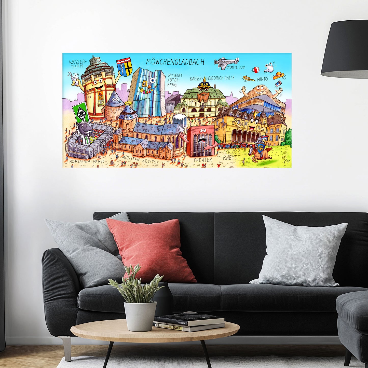 Jacques Tilly Mönchengladbach Stadtbild auf Leinwand mit Holzrahmen über einem schwarzen Sofa mit einem roten und zwei grauen Kissen und einem runden Couchtisch aus Holz davor.