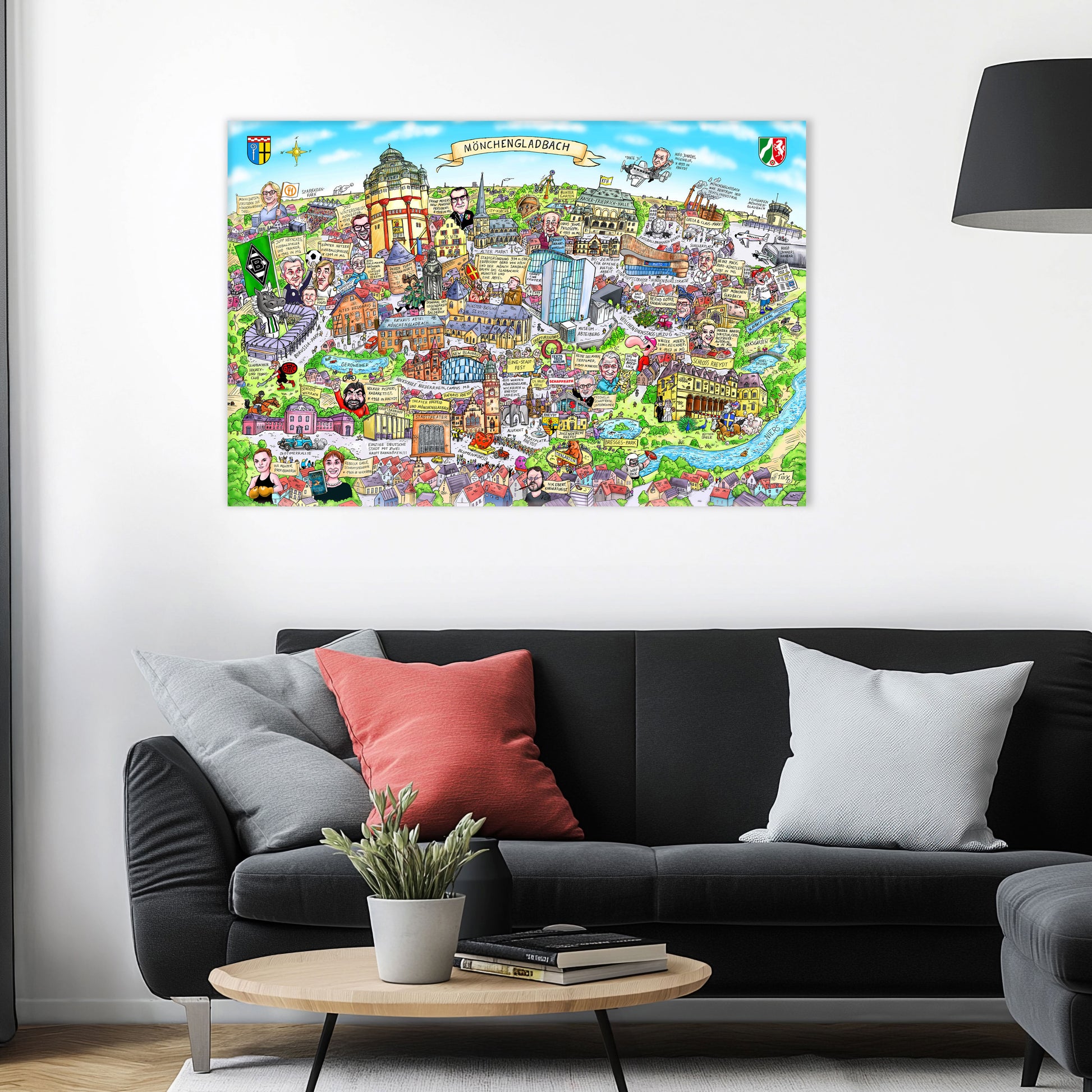 Jacques Tilly Mönchengladbach Stadtbild auf Leinwand mit Holzrahmen über einem schwarzen Sofa mit einem roten und zwei grauen Kissen und einem runden Couchtisch aus Holz davor.