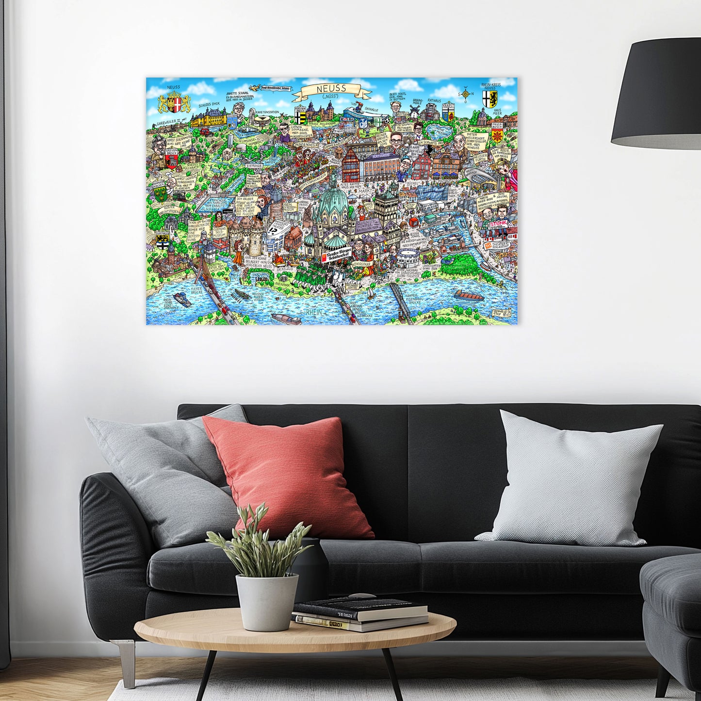 Jacques Tilly Neuss Stadtbild Wimmelbild auf Leinwand mit Holzrahmen über einem schwarzen Sofa mit einem roten und zwei grauen Kissen und einem runden Couchtisch aus Holz davor.