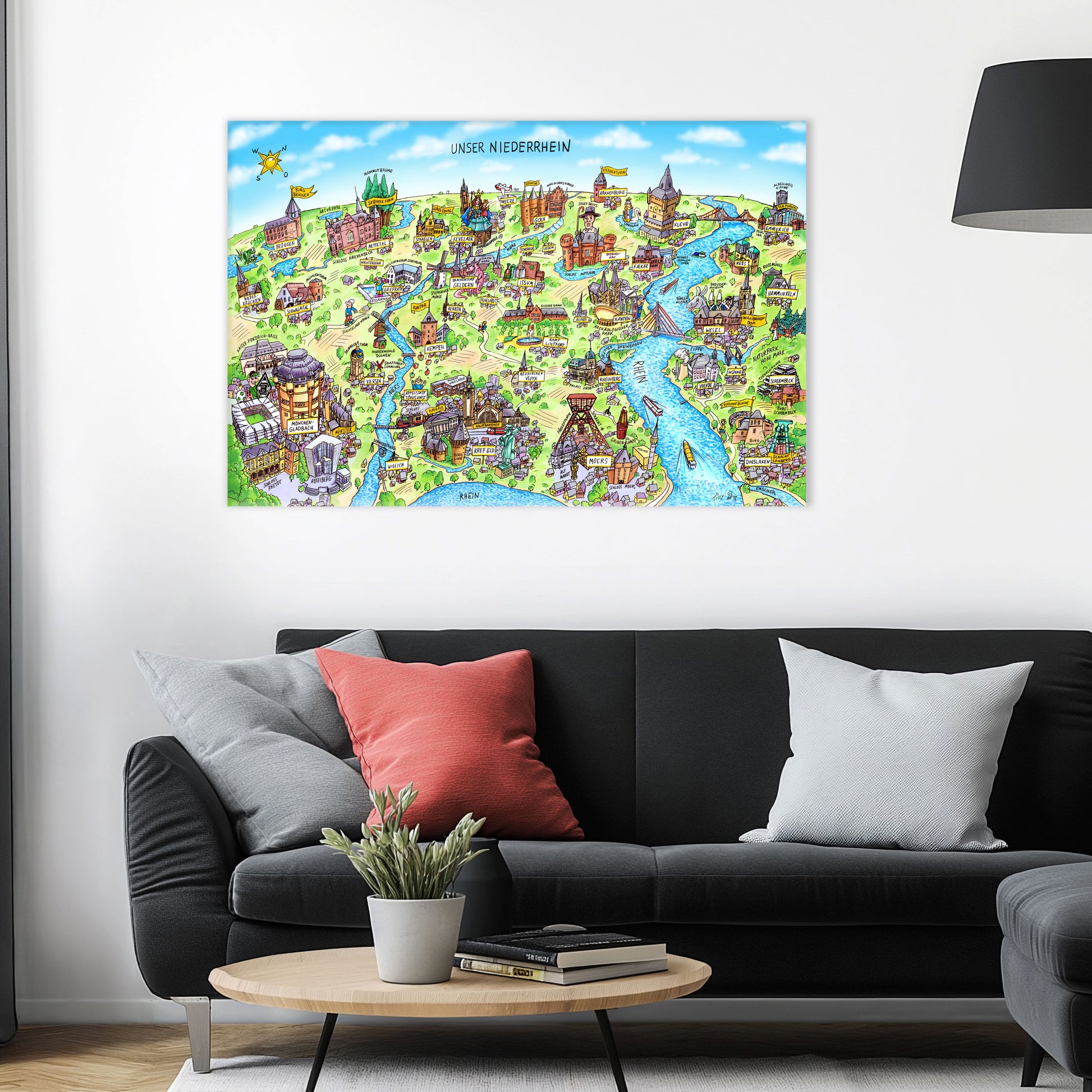 Jacques Tilly Niederrhein Wimmelbild als Acrylbild mit Alu-Rückrahmen über einem schwarzen Sofa mit einem roten und zwei grauen Kissen und einem runden Couchtisch aus Holz davor.